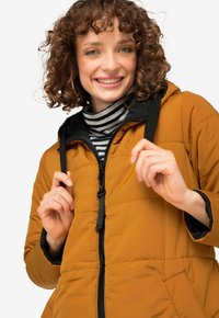 Mosterdgele gewatteerde jas met een zwarte capuchon en rits. Voorzien van elastische trekkoorden en een gestreept shirt eronder.