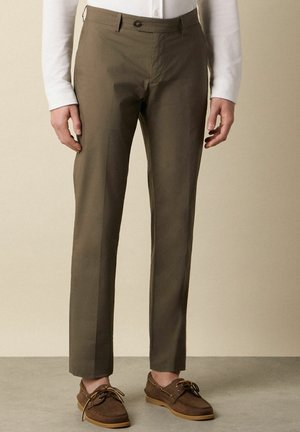 Uomo che indossa pantaloni eleganti verde oliva slim fit con mocassini in pelle marrone su uno sfondo neutro.