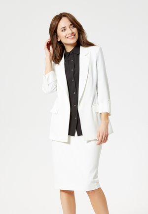 Vrouw glimlachend, draagt een wit blazer en rok met een zwart hemd met knopen, haar haar aanrakend tegen een effen lichte achtergrond.
