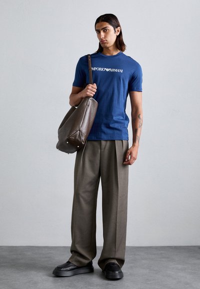 Mornarsko modra bombažna majica z logotipom "Emporio Armani", v kombinaciji s širokimi olivno zelenimi hlačami in rjavim usnjenim nahrbtnikom. Črne čevlje.
