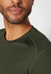 Zeleno športno majico s kratkimi rokavi, ki ima okroglo izrezanje in črn logotip "BORG" na rami. Gladka tekstura, prilegajoča zasnova.