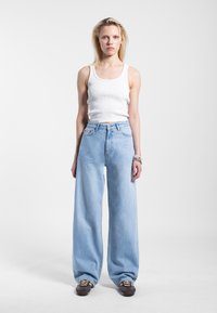 Lichtblauwe jeans met brede pijpen en een hoge taille, gecombineerd met een aansluitend wit geribbeld tanktop. Het model draagt bruine schoenen met veters en armbanden.
