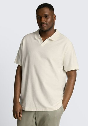 Mężczyzna stoi z jedną ręką w kieszeni, ubrany w kremową koszulkę polo z krótkim rękawem i jasne spodnie na jednolitym tle.