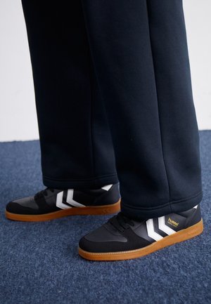 Zapatillas blancas y negras con suelas de goma, usadas con pantalones negros, de pie sobre una alfombra azul contra un fondo blanco liso.