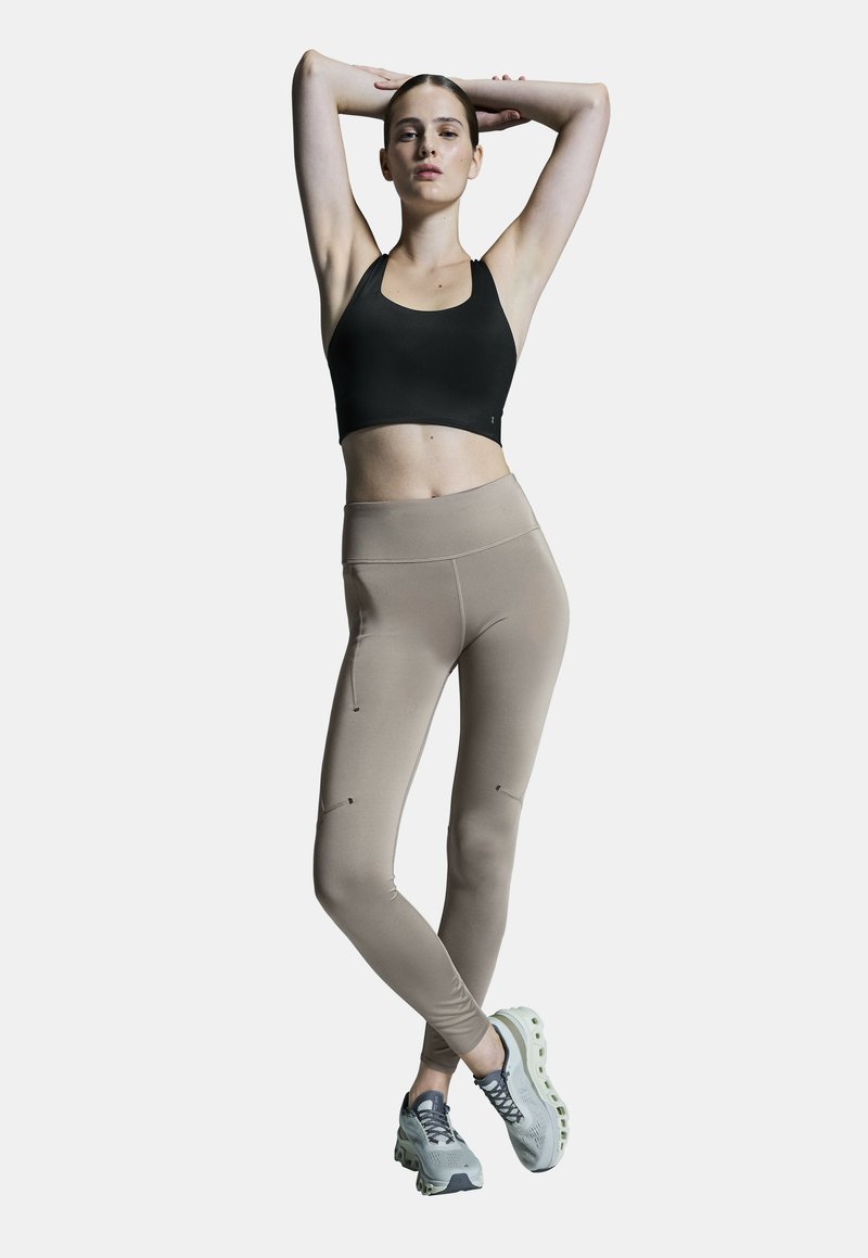 Schwarer Sport-BH und beigefarbene hochtaillierte Leggings mit Seitennähten und Akzenten. Das Modell trägt hellgraue Sportschuhe mit einer gepolsterten Sohle.