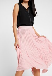 Musta hihaton paita, jossa on V-muotoinen kaula-aukko ja vaaleanpunainen laskostettu midi-hame. Hameessa on tiukka vyötärö ja virtaava, teksturoidu kangas.