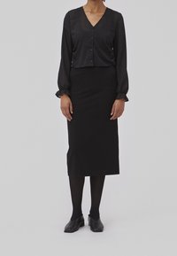 Blouse noire texturée à manches longues et fermeture à boutons, associée à une jupe crayon noire jusqu'aux genoux et des chaussures noires.