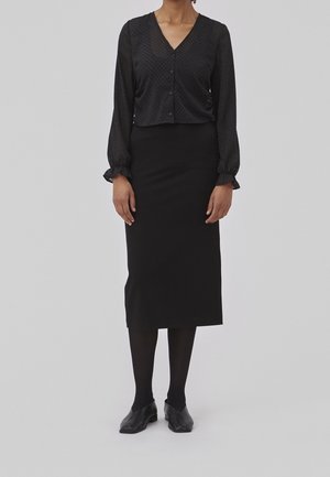 Blouse noire texturée à manches longues et fermeture à boutons, associée à une jupe crayon noire jusqu'aux genoux et des chaussures noires.