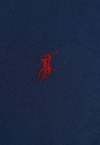 Námořnická modrá tkanina s malým červeným vyšitým logem pólisty. Textura je hladká s úzkou vazbou a design je minimalistický.