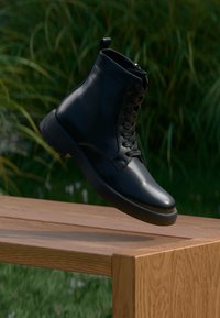 Bottine noire en cuir à lacets flottant au-dessus d'un banc en bois avec de l'herbe verte et des plantes en arrière-plan.