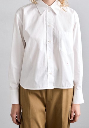 Persoon draagt een wit overhemd met knopen, een kraag en een borstzak, gecombineerd met bruine cargobroek, staand tegen een neutrale achtergrond.