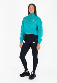 Felpa corta zip-up color teal con logo ricamato e leggings neri a vita alta con dettagli in rete e swoosh bianco; indossata con sneakers nere.