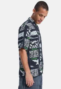 Μαύρο πουκάμισο με κοντά μανίκια και κουμπιά, διακοσμημένο με λευκά και πράσινα γραφικά μοτίβα, που περιλαμβάνουν αφηρημένα σχέδια και ανάγλυφες διακριτικές επιγραφές με το λογότυπο.