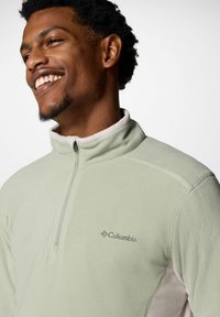 Pullover in pile verde chiaro con colletto a mezza zip, tessuto morbido e logo Columbia marrone. Presenta un colletto interno bianco a contrasto.