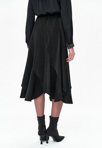 Czarna, błyszcząca asymetryczna spódnica z rozkloszowanym dołem, wyposażona w teksturowany pas i warstwowy design. Zestawiona ze spiczastymi botkami za kostkę.