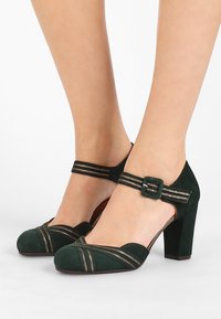 Chaussures à talons hauts en suède vert avec un orteil arrondi et une sangle pour la cheville. Accents décoratifs à rayures dorées à l'avant et sur la sangle. Design à talon carré.