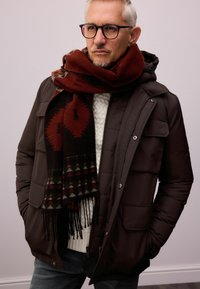 Braune Steppjacke mit vorderen Taschen, weißer Kabelstrickpullover und ein dunkelroter gemusterter Schal mit geometrischen Designs und Fransen.