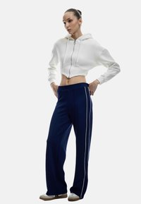 Eine gekürzte weiße Kapuzenjacke mit Reißverschluss, kombiniert mit navyblauen weiten Hosen mit weißen Seitenstreifen. Das Modell trägt lässige Turnschuhe.