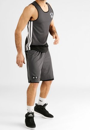 Mężczyzna w czarno-szortach do koszykówki i bezrękawniku, w czarnych butach Adidas i białych skarpetkach Adidas na białym tle.