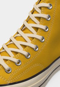 Baskets montantes en toile jaune avec des lacets blancs passés à travers des œillets argentés et une semelle en caoutchouc blanche avec une bande noire.