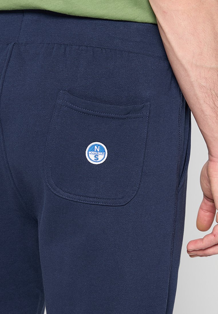 I pantaloni della tuta blu navy presentano una tasca posteriore con un logo bianco tondo con la scritta "NORTH SAILS". Sono realizzati in un tessuto morbido con una superficie liscia.