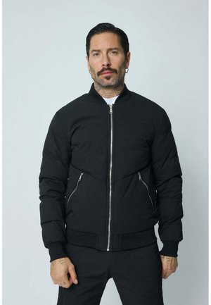 Veste en duvet noire avec fermeture éclair à l'avant, dotée de deux poches latérales zippées, de poignets côtelés et d'un col rond. Texture lisse et design rembourré.