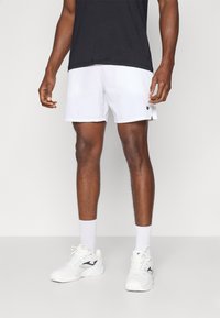 Shorts de sport blancs avec une taille élastique et une fente sur le côté, associés à des baskets blanches et des chaussettes blanches jusqu'aux genoux. Un t-shirt noir est porté.