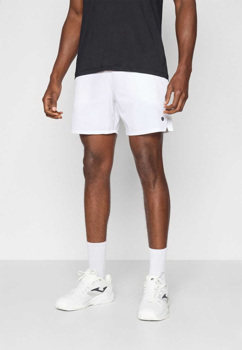 Shorts de sport blancs avec une taille élastique et une fente sur le côté, associés à des baskets blanches et des chaussettes blanches jusqu'aux genoux. Un t-shirt noir est porté.