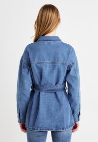 Chaqueta de mezclilla en azul claro con cintura ceñida. Presenta un collar en punta, mangas largas y un corte relajado con detalles de costura visibles.