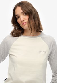Jonge vrouw met schouderlang bruin haar, gekleed in een creme- en lichtgrijze Superdry raglan mouwen shirt, tegen een effen witte achtergrond.