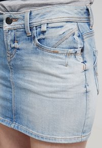 Ljusblå denimkjol med frontfickor, bältesloops och bruna sömmar, som bärs på höfterna.