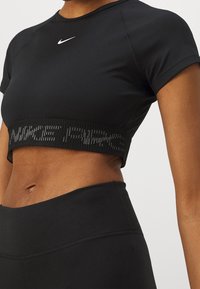 Musta lyhyt tekninen paita joustavasta kankaasta. Siinä on pyöreä kaula-aukko ja leveä nauha, jossa on ristiriidassa oleva "NIKE"-teksti.