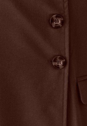 Primer plano de tela marrón con dos botones redondos y brillantes de cuatro agujeros y una vista parcial de un bolsillo con solapa en el lado derecho.