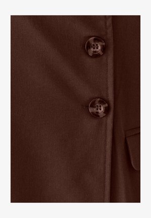 Primer plano de tela marrón con dos botones redondos y brillantes de cuatro agujeros y una vista parcial de un bolsillo con solapa en el lado derecho.