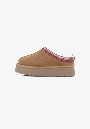 Zapato slip-on de gamuza color tostado con una gruesa suela de plataforma beige y ribete tejido acentuado en rojo a lo largo del borde superior. Cuenta con una lengüeta en la parte trasera.
