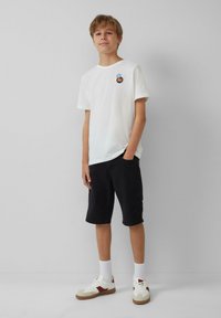 Witte katoenen t-shirt met een kleine graphic op de linkerborst, gecombineerd met zwarte shorts en witte sneakers met rode accenten.