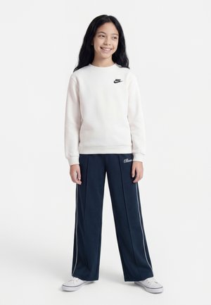 Fille aux longs cheveux noirs portant un sweat-shirt blanc Nike, un pantalon large bleu marine avec un liseré blanc, et des baskets blanches, debout et souriante.