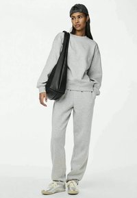 Young person wearing gray sweatshirt and sweatpants, silver sneakers, black cap, and carrying large black shoulder bag, standing against white background.