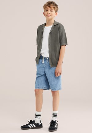 Jeune garçon debout portant un cardigan gris à manches courtes, un t-shirt blanc, un short en jean bleu, des chaussettes blanches avec des rayures noires et des baskets noires.