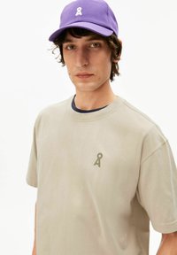 Beige katoenen T-shirt met een ronde hals en een klein olijfgroen logo. Gecombineerd met een paarse pet met een wit logo aan de voorkant.