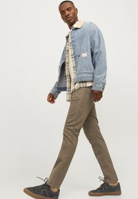 Uomo che cammina in giacca di denim blu chiaro, camicia a quadri, pantaloni khaki e sneakers nere con suole in gomma, su uno sfondo chiaro e semplice.
