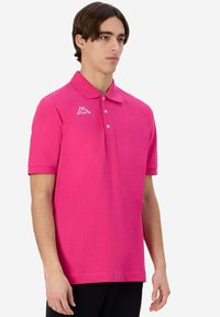 Maglietta polo rosa con colletto, maniche corte e patta con tre bottoni. Presenta un logo bianco sul lato sinistro del petto e una texture liscia.