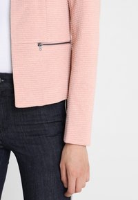 Veste rose pâle texturée au design ajusté, avec une poche zippée et des manches longues, associée à un jean foncé.