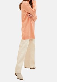 Hellorange gestrickter Pullover mit geripptem Saum, kombiniert mit cremefarbenen, weit geschnittenen Hosen und beigen Ankle Boots. Weiche Textur und lockere Passform.