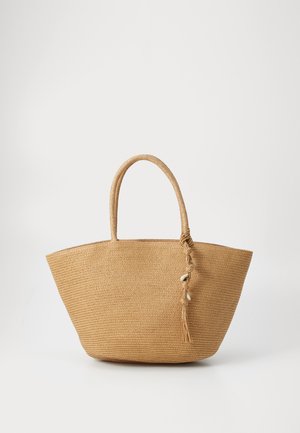 Bolso de paja trenzada en beige, con dos asas redondas y un detalle de borla con conchas. Base ancha y diseño de apertura superior.