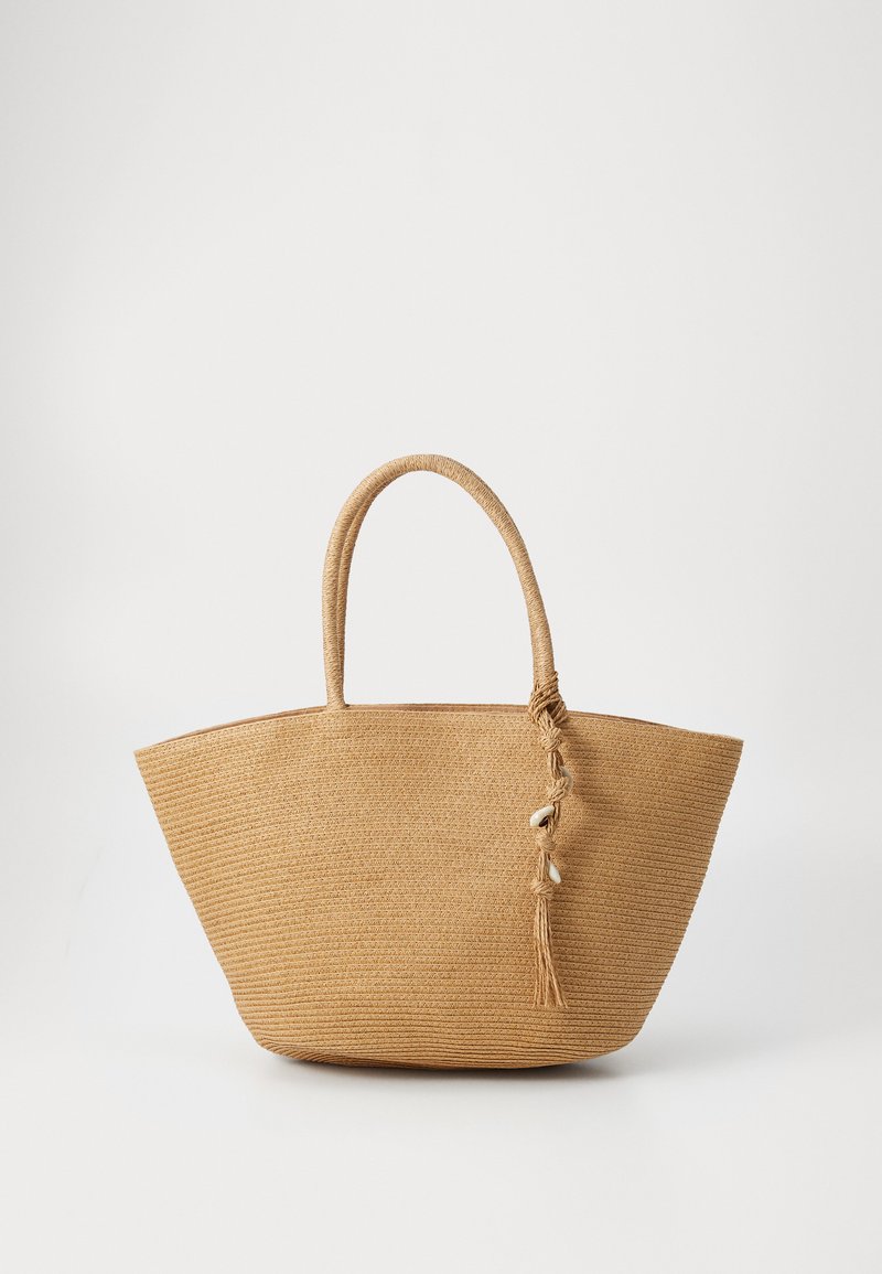 Woven straw tote bag v bežni barvi, ki ima dve okrogli ročki in detajl s frufruji ter školjkami. Široka osnova in odprt zgornji del.
