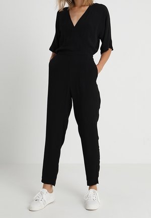 Kobieta w czarnym kombinezonie z krótkim rękawem i dekoltem w serek, z kieszeniami oraz białymi trampkami na sznurówki, stojąca na jednolitym jasnym tle.