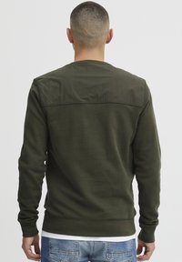 Olive green sweatshirt with a textured back panel, crew neck, long sleeves, and ribbed cuffs, layered over a white shirt.