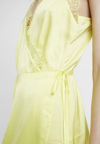 Keltainen satiinimekko pitsiyksityiskohdilla, jossa on kietaisusuunnittelu, säädettävä sidonta vyötäröllä ja sileä, herkkä tekstuuri.
