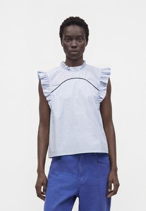 Femme portant un chemisier sans manches bleu clair à rayures avec des volants aux épaules et au col, associé à un pantalon bleu taille haute.